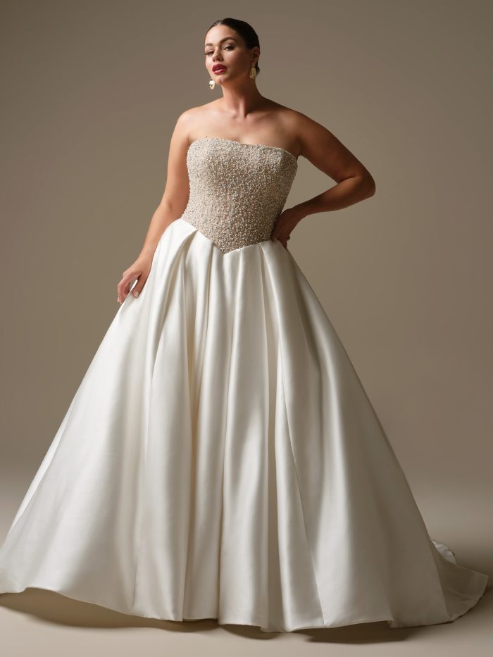 Model wearing Dmara by Sottero and Midgley, a strapless Cartago satin ball gown wedding dress with a beaded sheer bodice, straight neckline, and full skirt with pockets.