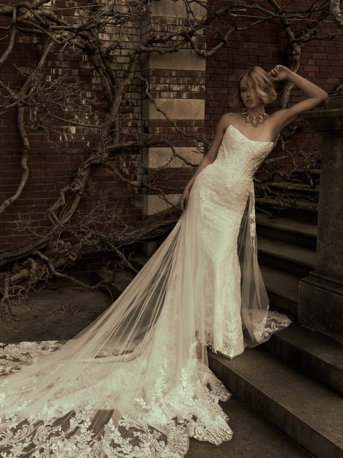 Bride wearing the Eirawen fit-and-flare wedding dress by Sottero and Midgley, featuring a strapless beaded lace bodice and dramatic sheer lace-trimmed train, standing against a brick wall with bare branches.
