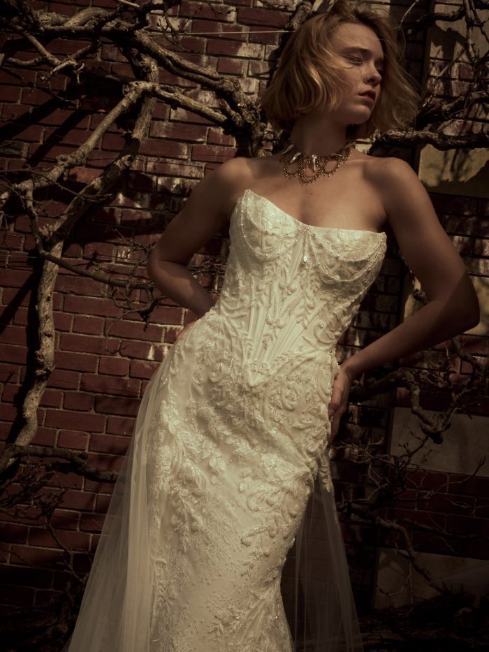 Close-up of a bride wearing the Eirawen fit-and-flare wedding dress by Sottero and Midgley, showcasing the intricately beaded corset bodice with scalloped neckline, against a brick wall with bare branches.