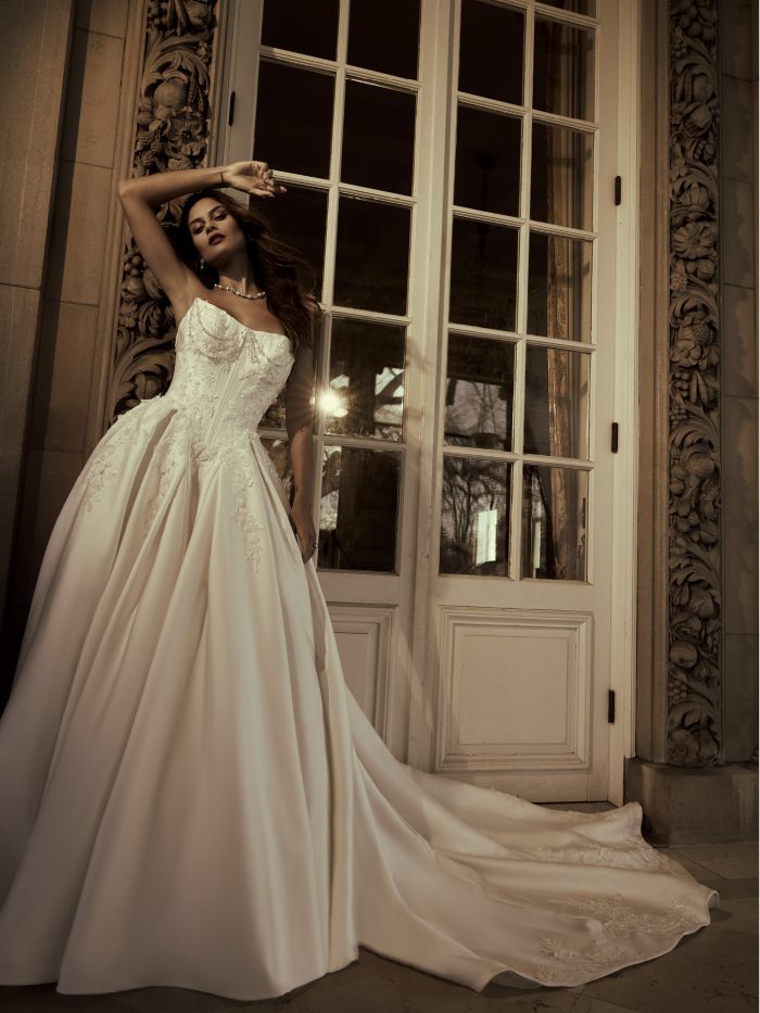 Bride wearing the Petra ballgown wedding dress by Sottero and Midgley, featuring a beaded strapless bodice with floral lace appliqués and a full satin skirt with train, standing before ornate carved doors.