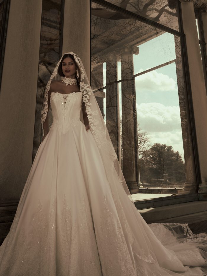 Bride wearing the Sandrine ballgown wedding dress by Sottero and Midgley, featuring a beaded strapless bodice and full satin skirt with lace hem detail, paired with a lace-trimmed cathedral veil, standing beside grand columns.