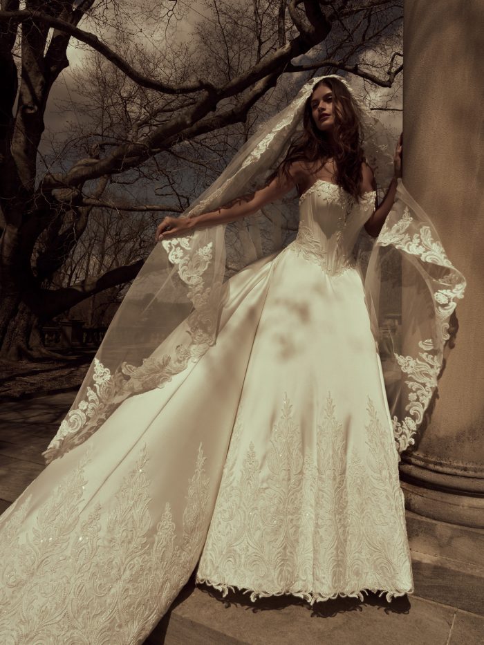 Bride wearing the Sandrine ballgown wedding dress by Sottero and Midgley, showcasing the beaded corset bodice and lace-appliquéd skirt, with a flowing lace-trimmed veil, standing outdoors beside stone columns and bare trees.