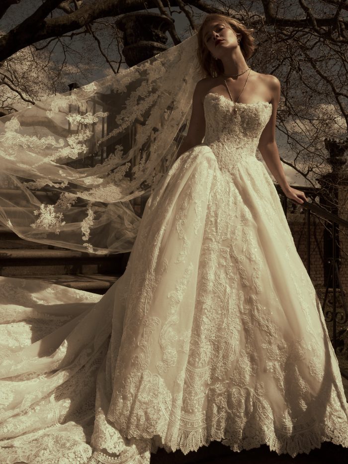 Bride wearing the Yesenia ballgown wedding dress by Sottero and Midgley, featuring a strapless beaded lace bodice and full lace-appliquéd skirt, paired with a flowing lace-trimmed cathedral veil, standing outdoors among bare trees.