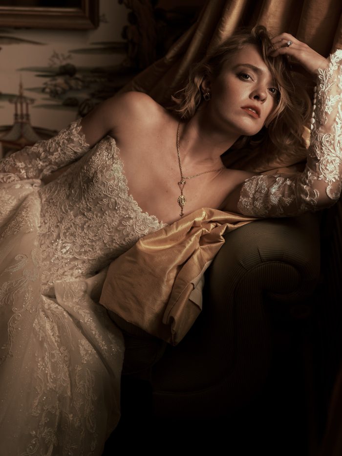 Close-up of a bride wearing the Yesenia ballgown wedding dress by Sottero and Midgley, showcasing the detachable lace sleeves and beaded lace bodice, reclining in an ornate interior setting.