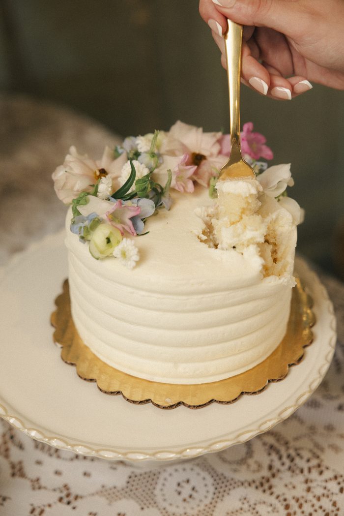 Small white wedding cake with smooth buttercream frosting and fresh pastel flowers, styled on a vintage lace-covered table as part of a romantic boho wedding dessert display.