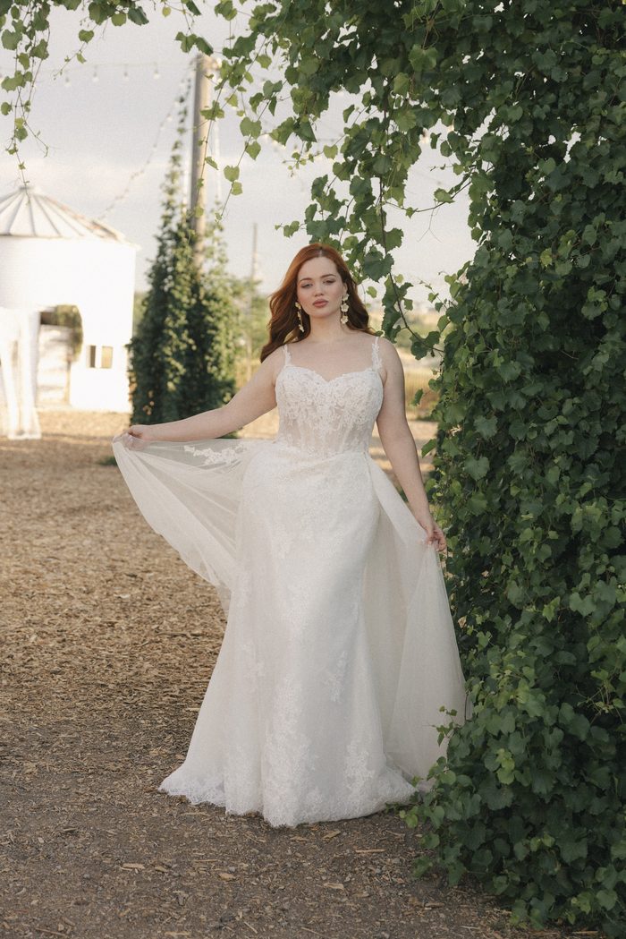 Full-length view of the Esmeralda Lane boho floral wedding dress with cascading floral lace, fitted silhouette, and soft tulle overskirt framed by lush green vines.