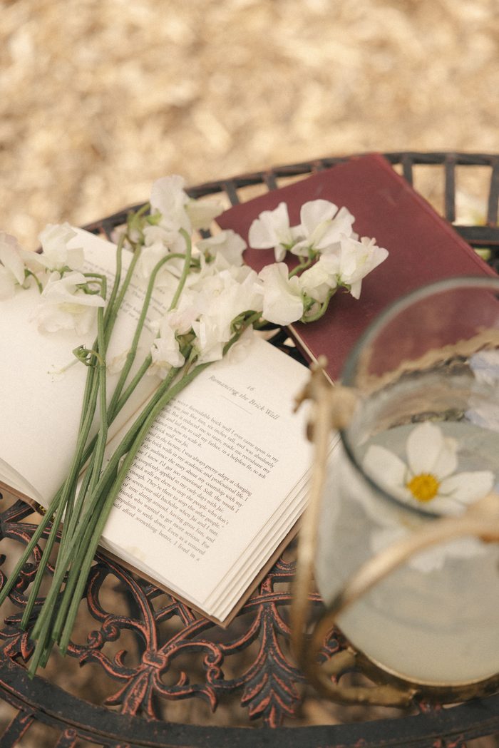 Vintage-inspired boho wedding detail featuring delicate white sweet peas resting on an open book beside a floating daisy in a glass bowl, creating a romantic garden-inspired tablescape.