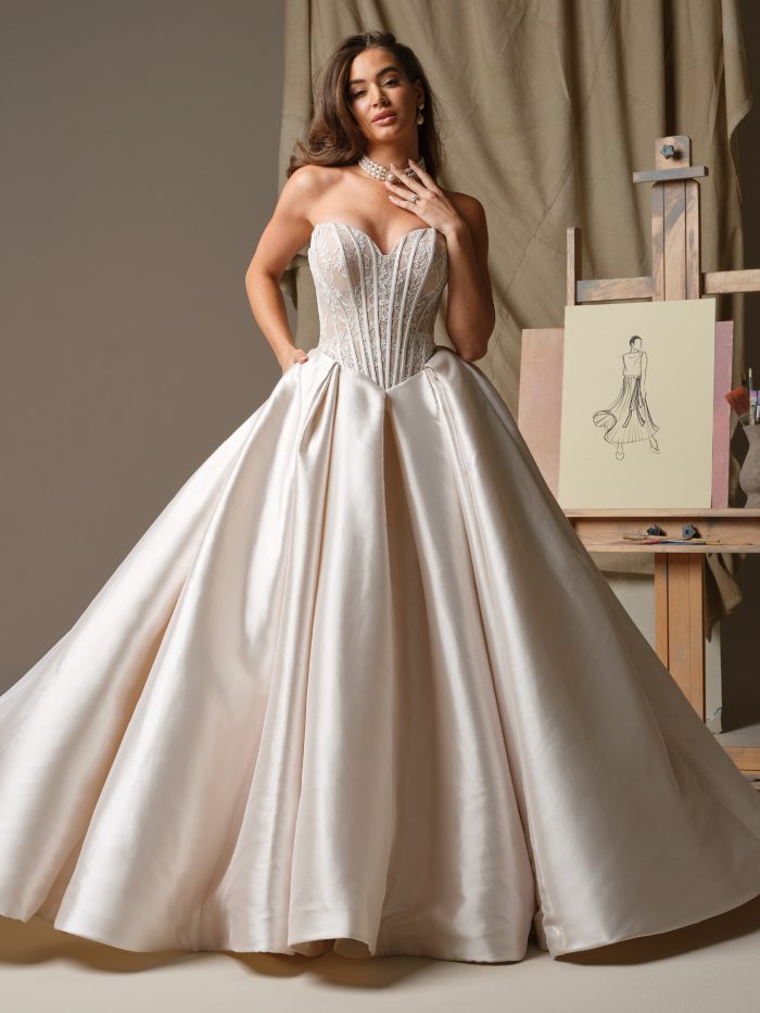 Ashland by Maggie Sottero. A glamorous strapless ballgown wedding dress featuring a sheer lace corset bodice with exposed boning and a full satin skirt with pleats, styled with pearl jewelry in an artistic studio setting.