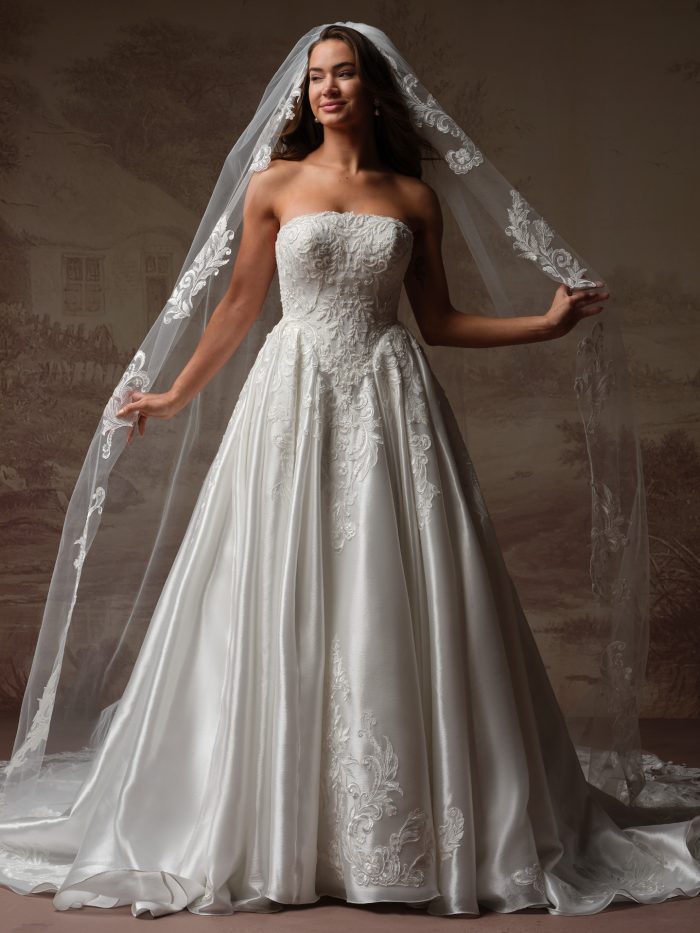 Full-length view of Maggie Sottero Dahlia ballgown with a strapless lace bodice, full satin skirt, and a long veil with embroidered lace edging.