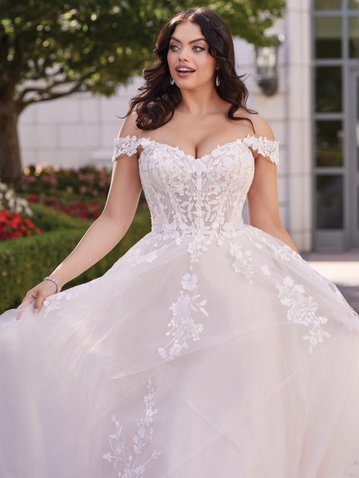 Bride wearing Harlem by Maggie Sottero, an off-the-shoulder ball gown wedding dress featuring a sweetheart neckline, sheer lace bodice with sequin floral appliqués, and a soft tulle skirt.