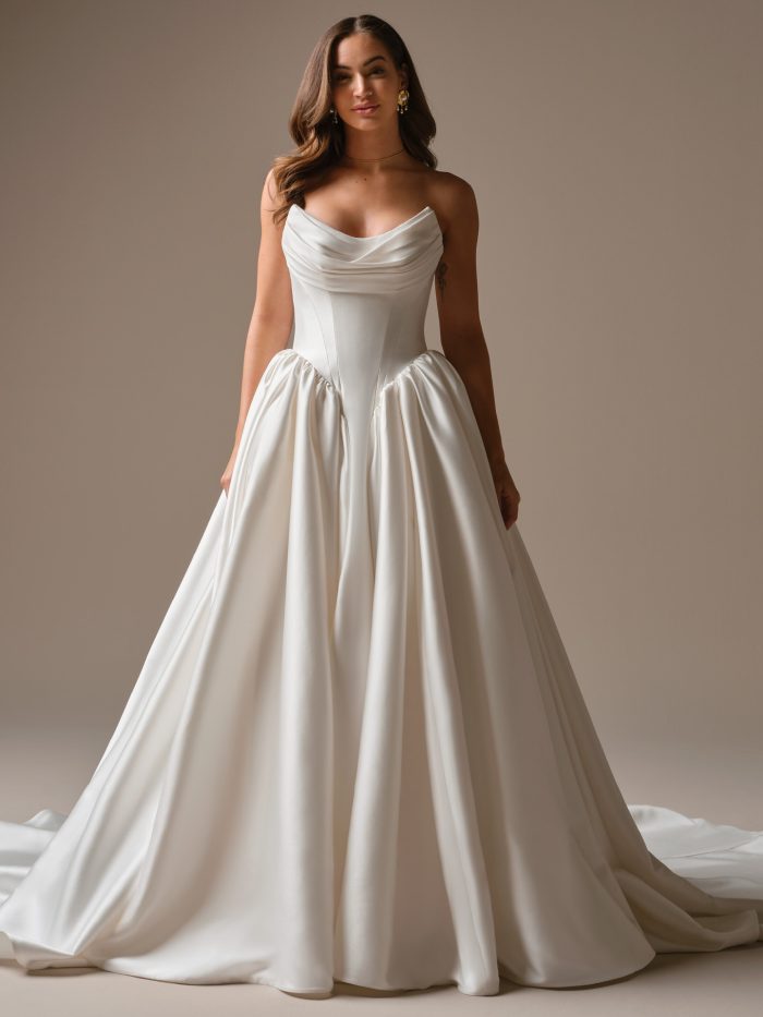 A modern satin ball gown wedding dress with a soft draped cowl neckline and structured bodice that cinches into a basque-inspired waist, flowing into a full, voluminous skirt.