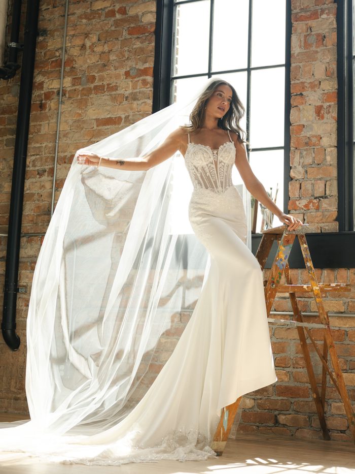 Bride wearing India by Maggie Sottero, a sheath wedding dress with a beaded illusion bodice, exposed boning, and lace motifs, paired with a long cathedral veil, shown in ivory.