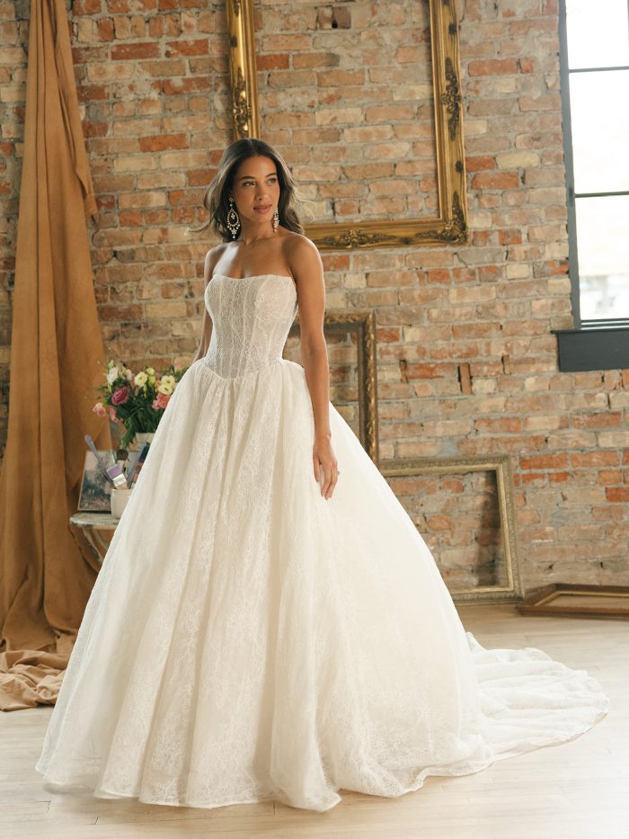 Strapless Chantilly lace ballgown wedding dress with sheer organza bodice and exposed boning, full skirt, and extended lace train styled in a rustic brick loft setting.