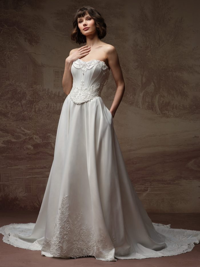 Full-length view of Maggie Sottero Seattle ballgown wedding dress with a strapless corset bodice, lace appliqué, pearl-draped neckline, and a full satin skirt with embroidered hem and train.