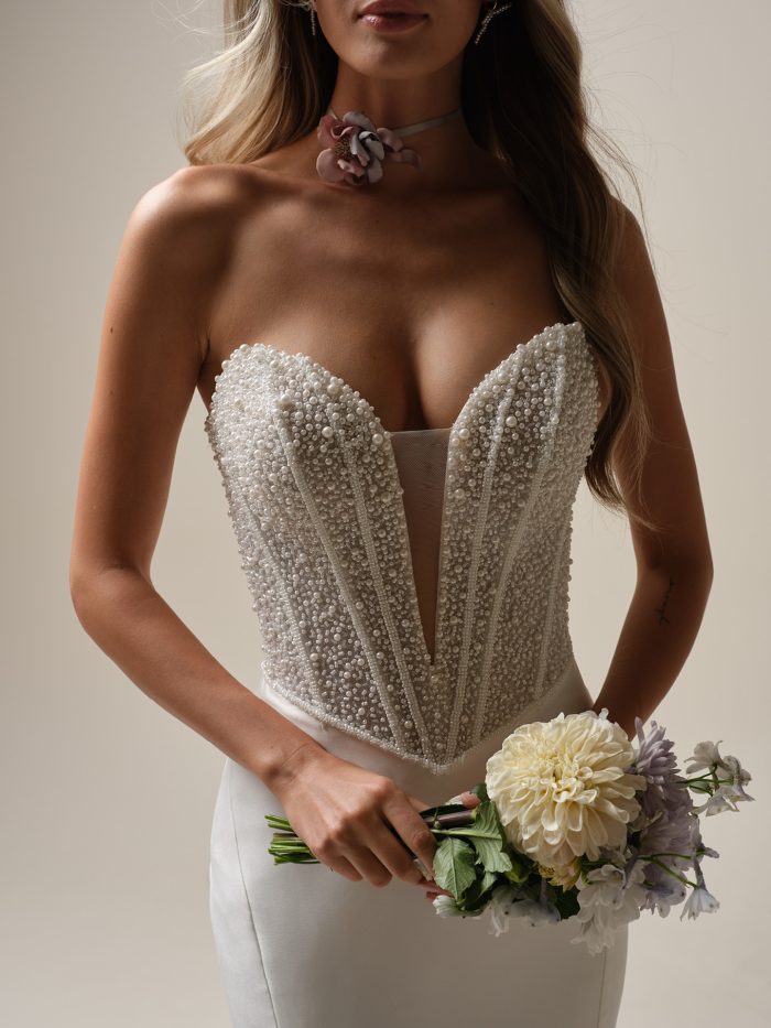Tina Marie by Maggie Sottero — close-up of a plunging strapless corset bodice adorned with pearl and bead detailing, paired with a sleek satin skirt and styled with a floral choker and soft bouquet in a studio setting.