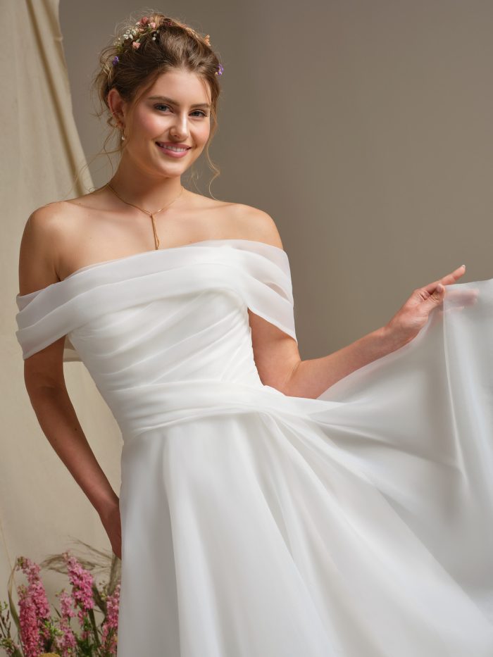 Bride in Dezi by Rebecca Ingram showcasing the off-the-shoulder Marquice organza neckline, pleated bodice detail, and airy full skirt with a soft sweeping hem.
