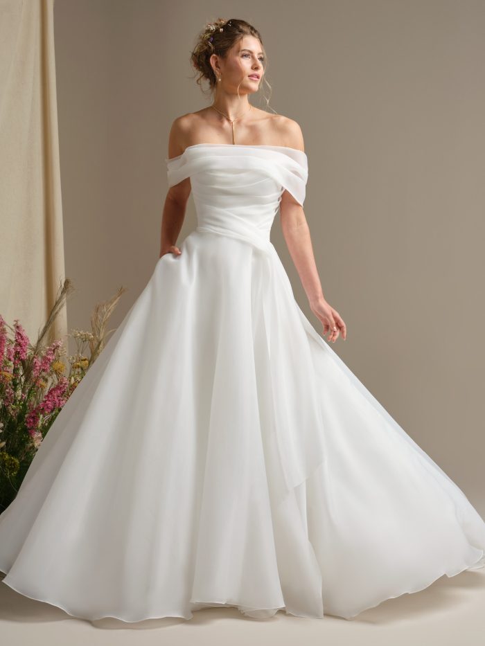Bride wearing Dezi by Rebecca Ingram, an off-the-shoulder organza ballgown wedding dress featuring an asymmetrically pleated bodice, soft draped neckline, and flowing A-line skirt.