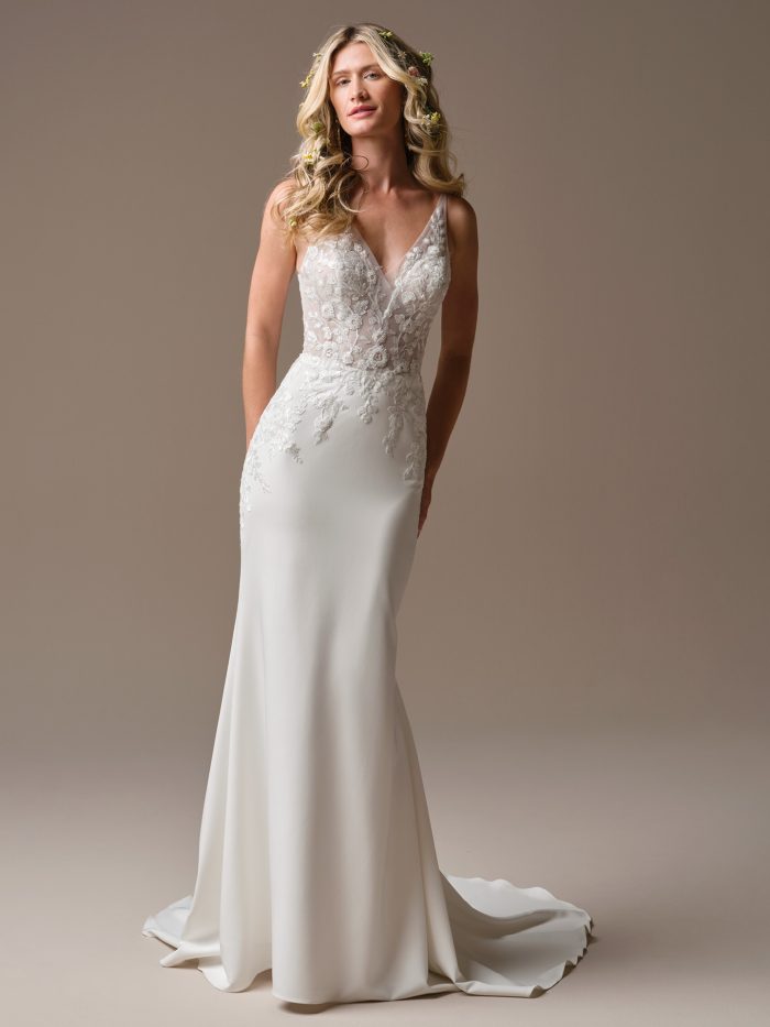 Bride wearing a modern sheath wedding dress with a deep V-neckline and illusion lace bodice. The gown features delicate floral lace appliqués that cascade onto the crepe skirt, creating a timeless and effortlessly elegant look.