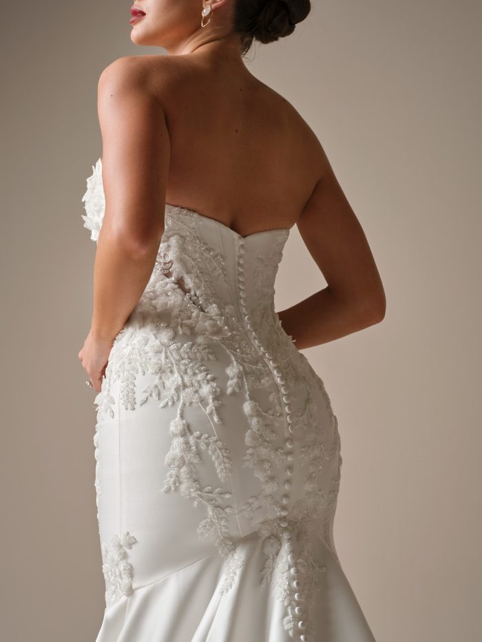 Alt text: “Back view of Sottero and Midgley Burke satin fit-and-flare wedding dress featuring beaded lace appliqués, corset bodice, and fabric-covered buttons trailing down the train.”