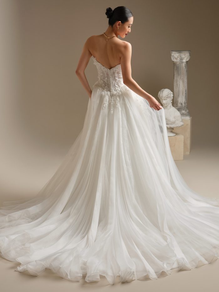 Back view of Jayna by Sottero and Midgley showcasing the illusion bodice with floral appliqués, covered button closure, basque waist, and flowing pleated glitter tulle train.