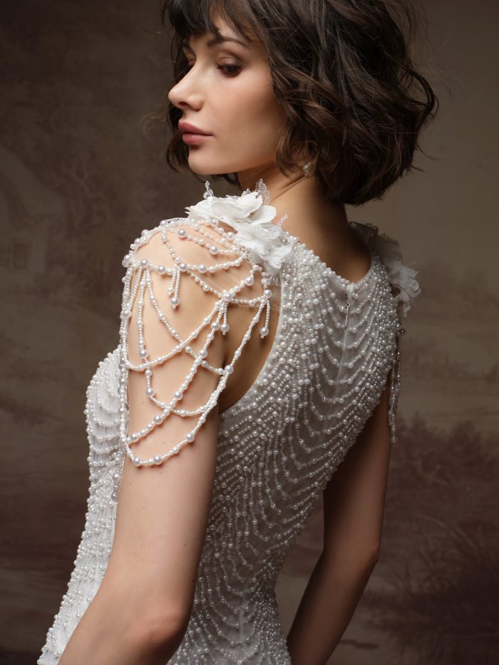 Close-up of Maggie Sottero Olata wedding gown featuring intricate pearl-beaded draping across the open shoulder and back, with delicate floral appliqué at the neckline and a textured, fully beaded bodice.