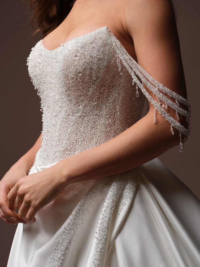 Close-up of Sottero and Midgley Rumi wedding gown featuring a strapless, heavily beaded bodice with crystal embellishments and delicate draped bead strands at the sleeves.
