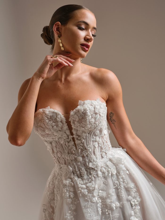 Close-up of a strapless A-line wedding dress with a deep sweetheart neckline and 3D floral lace appliqués. The bride poses gracefully, showcasing the gown’s intricate texture, sheer corset bodice, and soft tulle detailing.