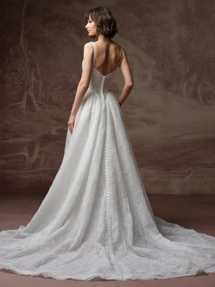 Back view of a bride wearing the Autumn ballgown wedding dress, showing a low V-back with thin straps, a row of covered buttons down the fitted lace bodice, and a full lace skirt sweeping into a train.