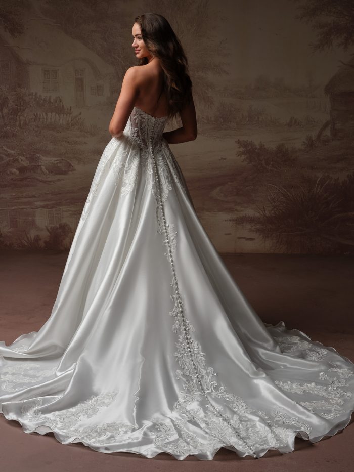 Back view of a bride wearing the Dahlia ballgown wedding dress, showing a sheer lace corset back with a row of covered buttons trailing down a lace-appliquéd satin skirt into a scalloped train.