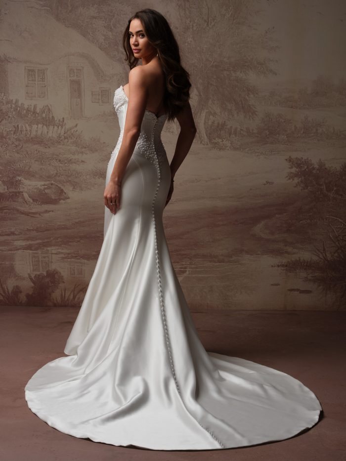 Back view of a bride wearing the Melissa Royale fit-and-flare wedding dress, showing a fitted satin bodice with pearl detailing at the waist, a row of covered buttons down the back, and a long train.