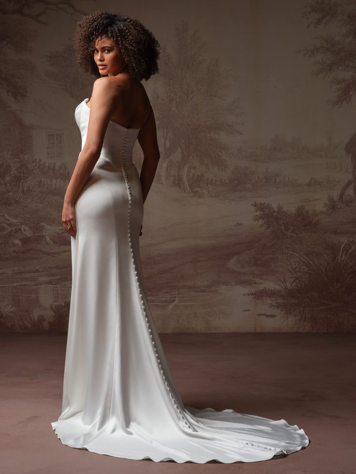 Back/side view of a bride wearing the Prestyn sheath wedding dress, showing a fitted strapless satin bodice with a row of covered buttons down the back and a flowing train.