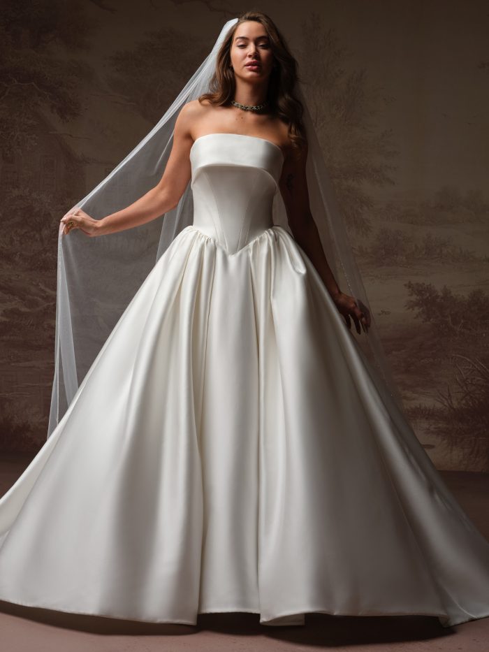 Front view of a bride wearing the Rane ballgown wedding dress in satin, featuring a strapless folded neckline, fitted bodice, and a full pleated ballgown skirt with train, paired with a sheer veil.