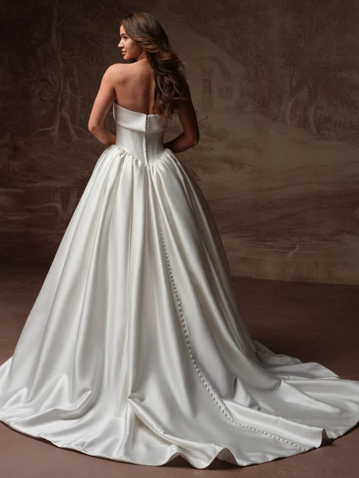 Back view of a bride wearing the Rane ballgown wedding dress, showing a strapless satin bodice with a row of covered buttons and a voluminous pleated skirt sweeping into a scalloped-edge train.