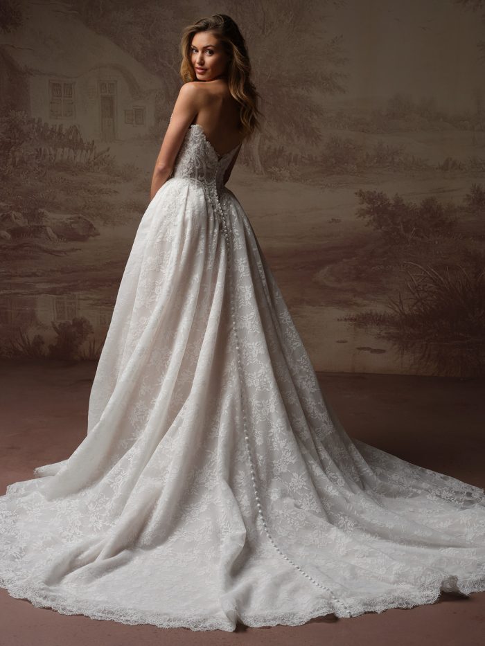 Back view of a bride wearing the Ryland ballgown wedding dress, showing a low lace back with a row of covered buttons down the fitted bodice and a voluminous lace skirt sweeping into a scalloped train.