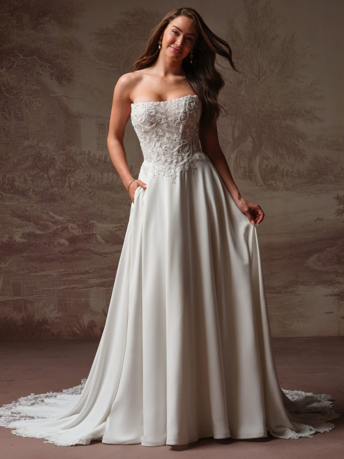 Front view of a bride wearing the Hazel A-line wedding dress, featuring a strapless lace-appliqued corset bodice and a flowing crepe skirt with pockets and a lace-trimmed train.