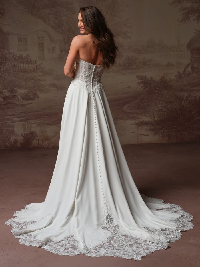 Back view of a bride wearing the Hazel A-line wedding dress, showing a sheer lace corset back with a row of covered buttons down the sweeping crepe skirt and scalloped lace-trimmed train.
