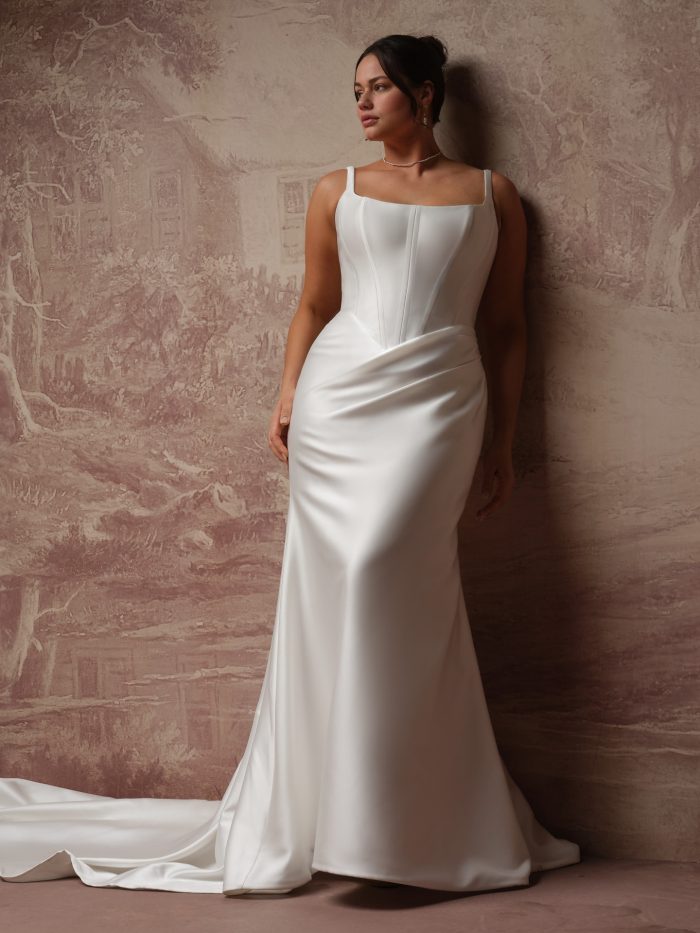 Plus-size bride wearing the Pamela fit-and-flare wedding dress in satin, featuring a fitted corset bodice with a squared neckline, slim straps, and a draped hip detail flowing into a sleek skirt with train.
