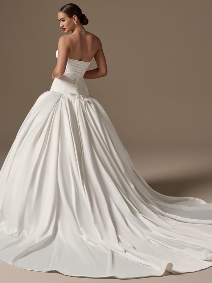Back view of a bride wearing the Adair wedding dress, showing a strapless bodice with corset lace-up back detail and a full pleated satin ballgown skirt sweeping into a train.