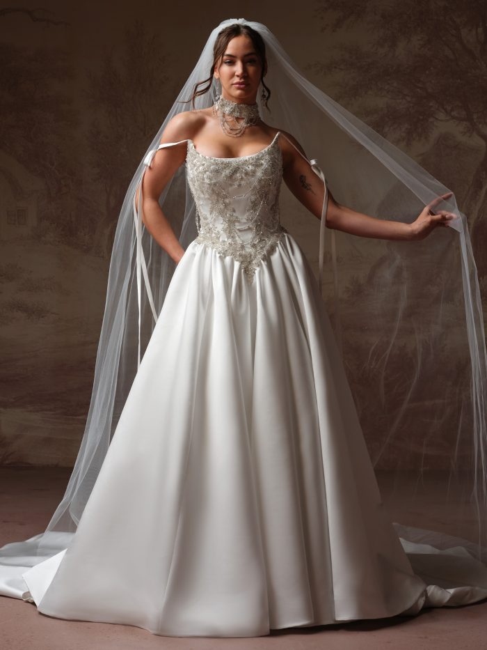 Bride wearing the Corazon veil from Maggie Sottero's Essentials Edit collection, a cathedral-length single-tier veil, paired with a satin ball gown featuring a beaded bodice and off-the-shoulder ribbon straps.
