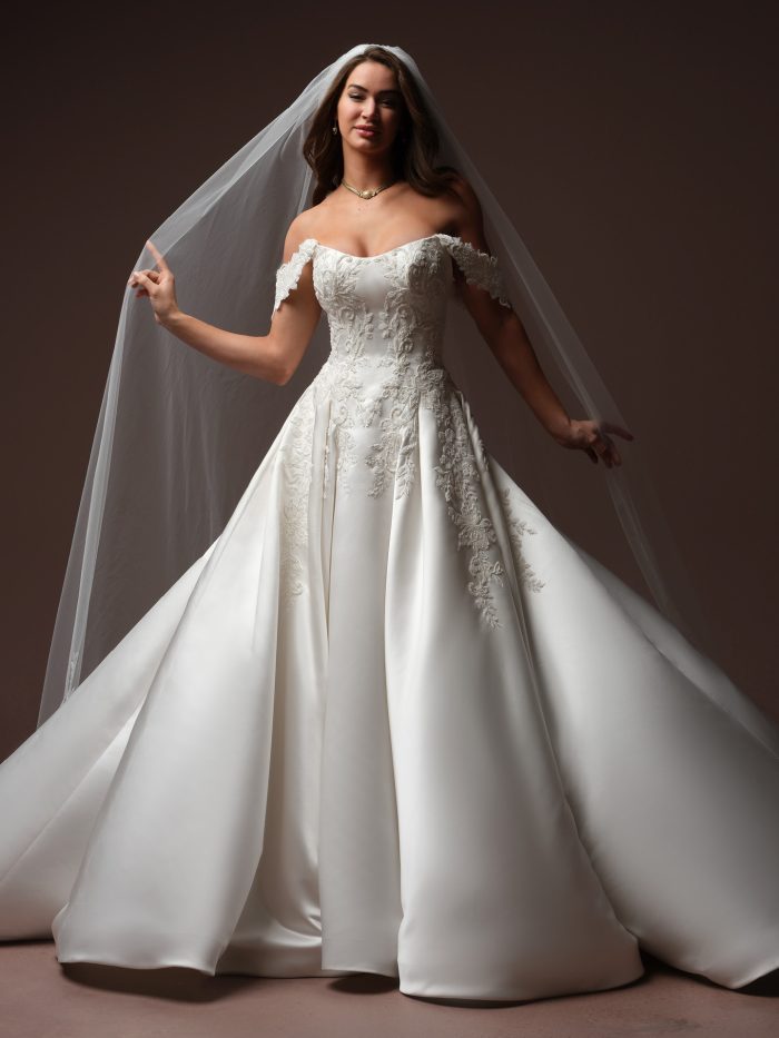 Bride wearing the Landis wedding dress by Maggie Sottero, featuring an off-the-shoulder lace bodice and full satin ball gown skirt, paired with a flowing cathedral veil.
