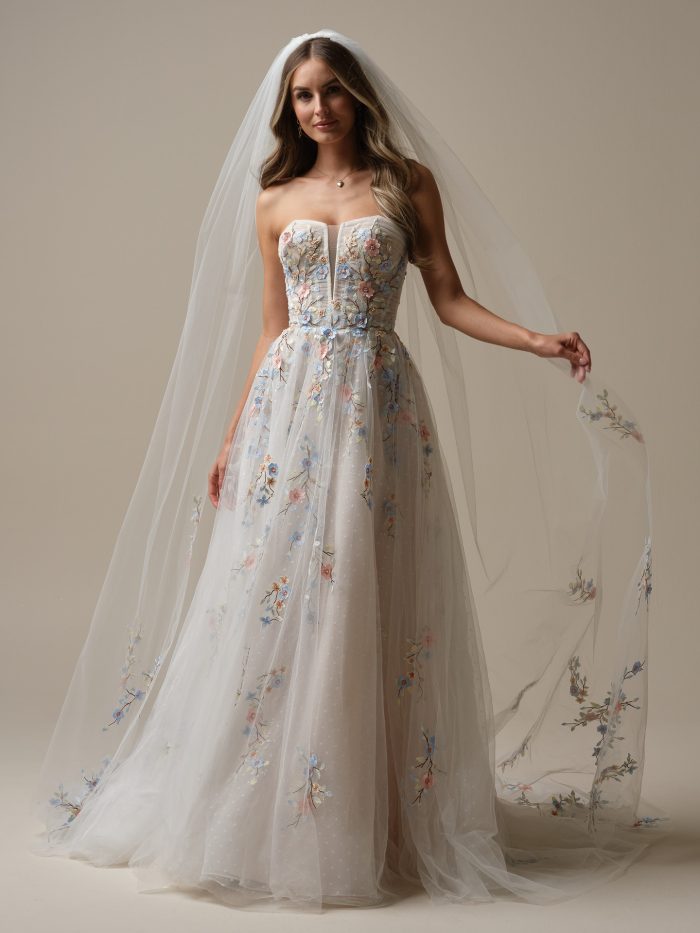 Bride wearing the Zephyrine A-line wedding dress by Maggie Sottero, featuring a strapless bodice with multicolor floral embroidery and a matching floral-embroidered veil in blue, pink, and gold tones.