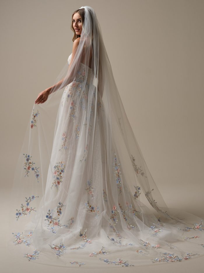 Back view of the Zephyrine A-line wedding dress by Maggie Sottero, showing the strapless silhouette with cascading multicolor floral embroidery on both the gown and flowing chapel-length veil.