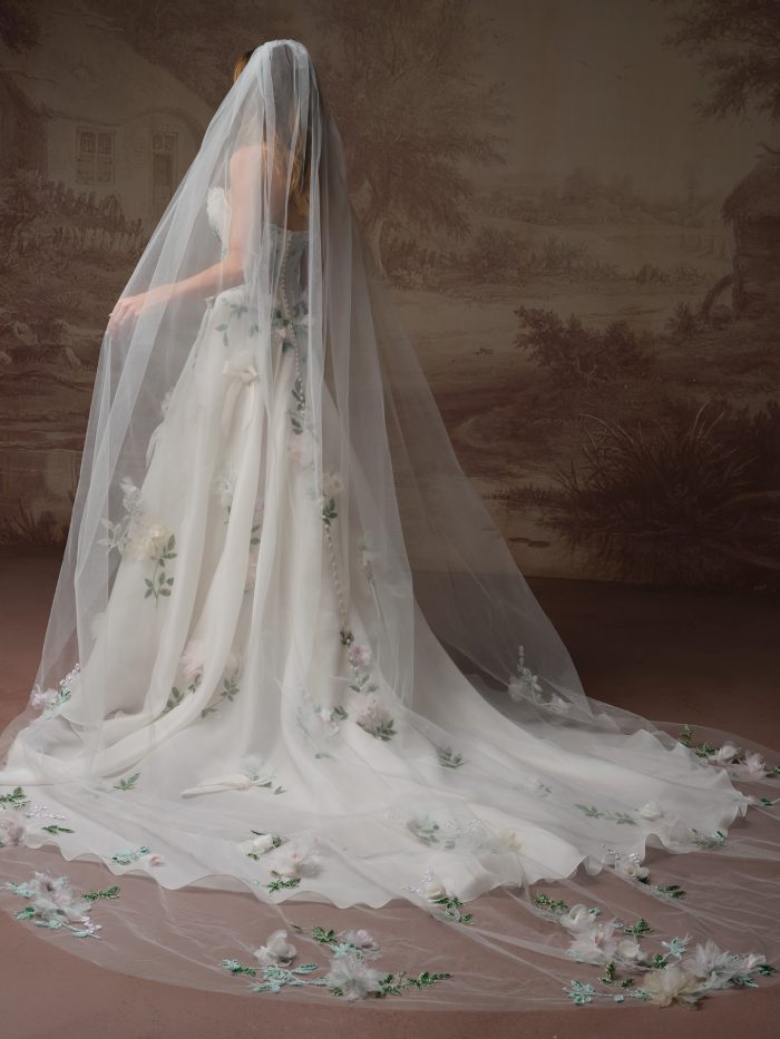 Back view of the Amaryllis wedding dress by Sottero and Midgley, showing the button-back bodice and flowing A-line skirt with cascading 3D florals, paired with a cathedral veil scattered with floral appliqué.
