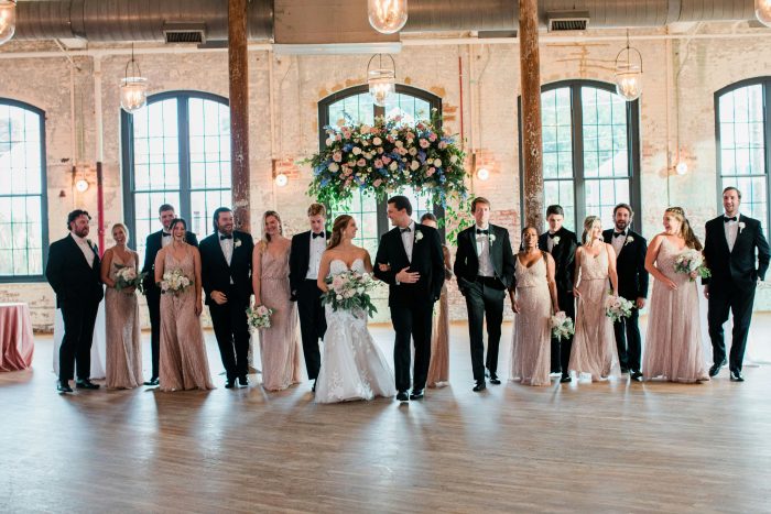 Bride wearing Hattie by Rebecca Ingram lace wedding dress walking with bridal party in an industrial wedding venue