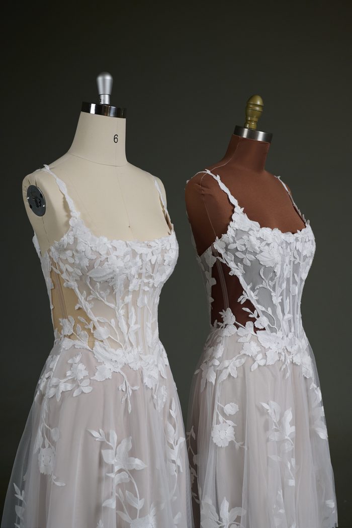 Two versions of the Natalie Marie by Maggie Sottero wedding gown are displayed side by side on dress forms, demonstrating different illusion mesh color options. Both feature a sheer corset bodice with exposed boning, a scoop neckline, floral lace appliqués, and a soft tulle skirt. The gown on the left showcases a lighter nude illusion mesh, while the gown on the right features a darker nude illusion mesh designed to better complement deeper skin tones while maintaining the gown’s delicate sheer effect.