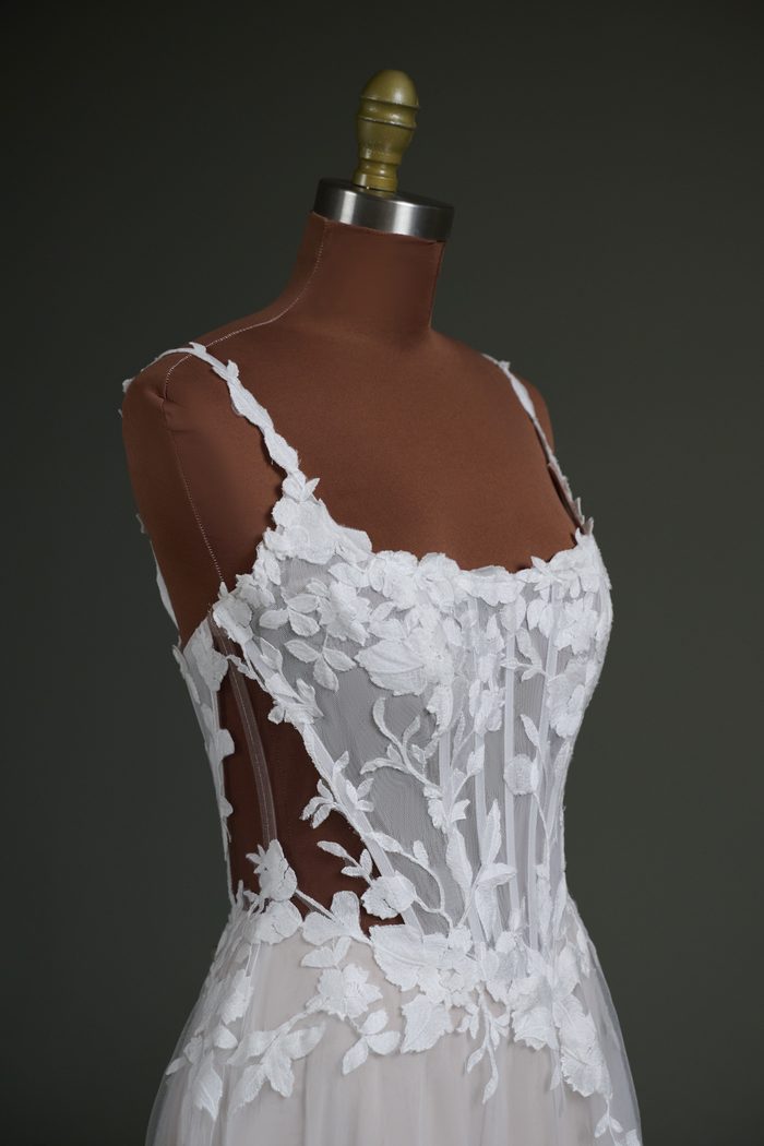 Close-up side view of the Natalie Marie by Maggie Sottero wedding gown displayed on a dress form with a dark nude mesh cover. The gown features a sheer corset bodice with exposed boning, a scoop neckline, and floral lace appliqués. The darker nude mesh highlights the illusion side panels and transparent construction, demonstrating a customization option designed to better complement deeper skin tones while maintaining the gown’s delicate lace detailing.