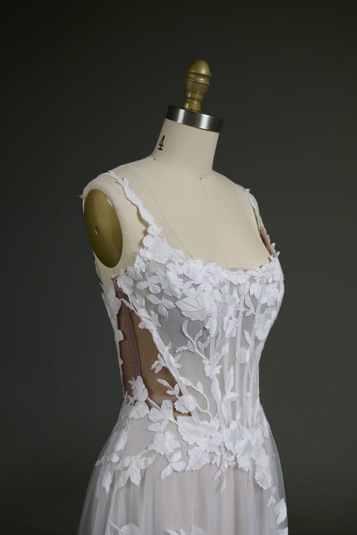 Close-up of the Natalie Marie by Maggie Sottero wedding gown bodice displayed on a dress form. The gown features a sheer corset bodice with visible boning, floral lace appliqués, and a scoop neckline. Darker nude illusion mesh is used in the side panels and bodice, demonstrating a customization option that provides a more seamless blend for deeper skin tones while preserving the gown’s transparent lace design.