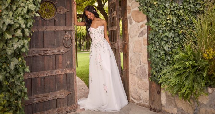Bride in the Blossom gown by Rebecca Ingram, a strapless A-line wedding dress with hand-painted pink floral appliqués, standing in a rustic wooden garden gate framed by ivy and stone walls.