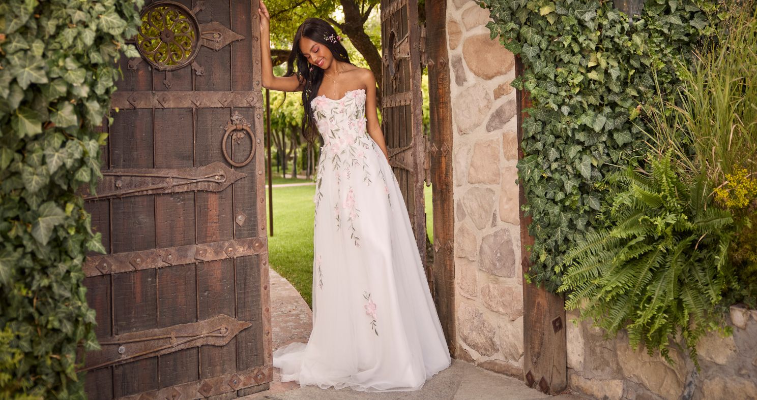 Bride in the Blossom gown by Rebecca Ingram, a strapless A-line wedding dress with hand-painted pink floral appliqués, standing in a rustic wooden garden gate framed by ivy and stone walls.