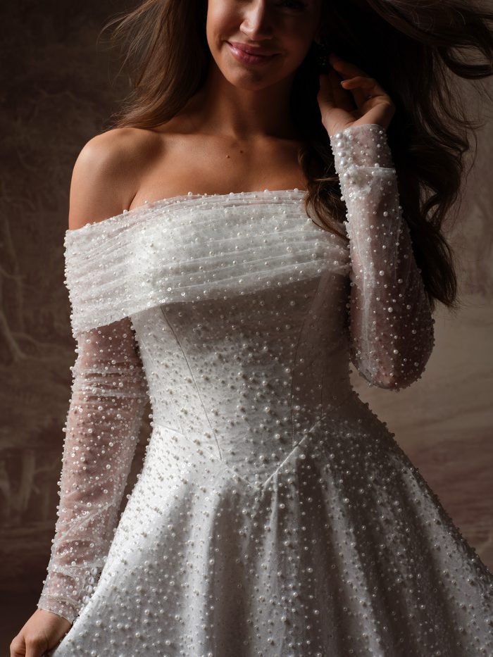 Maggie Sottero Elowyn Bridal Jacket, front detail of the pearl and sequin-embellished off-the-shoulder jacket with a folded portrait collar, structured corset bodice, and sheer long sleeves densely scattered with pearls of varying sizes.